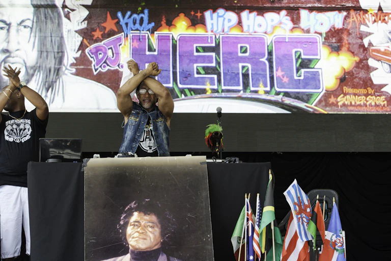 A rapper holds up both of his arms crossed while performing on stage. Behind him is a giant billboard that reads 'D.J. HERC' in graffiti.