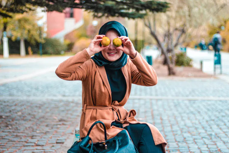 Woman in hijab smiles and holds two pieces of fruit over her eyes like binoculars.