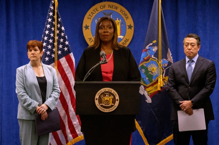 James, center, at a podium with two people flanking her. An American flag and New York State flag are behind them