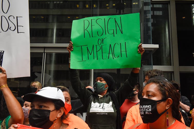 Crowd on the street, wearing face masks, with a woman holding a 'Resign or Impeach' sign