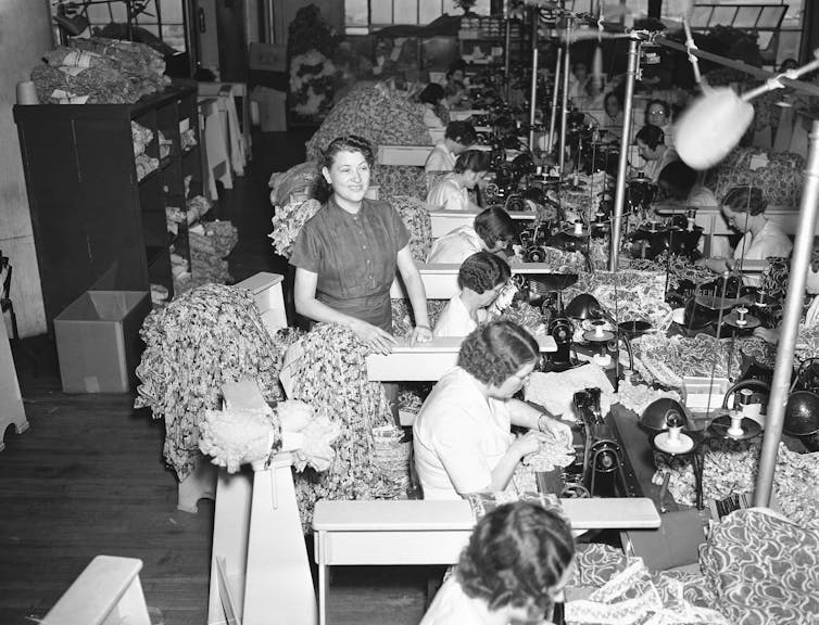A large room is full of women sitting at tables and using sewing machines to make garments, while a woman is standing, in 1937