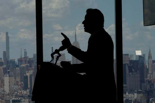 A dark image of Gov. Cuomo's profile while standing at a microphone and pointing with his left hand.