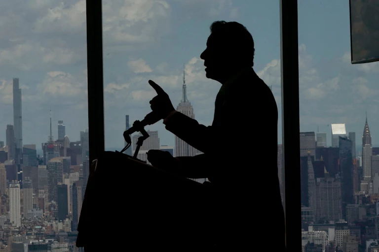 A dark image of Gov. Cuomo's profile while standing at a microphone and pointing with his left hand.