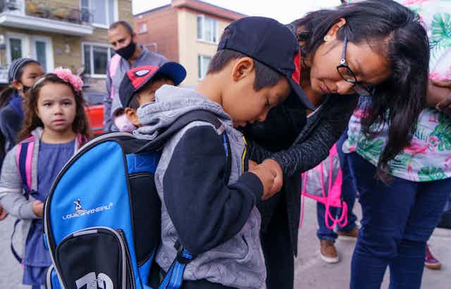 A mother helps her son in a backpack zip up a jacket.