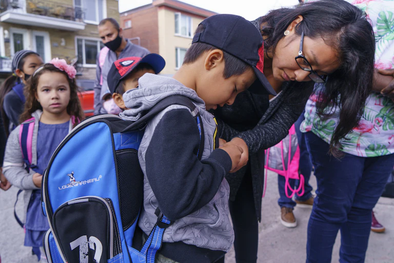 A mother helps her son in a backpack zip up a jacket.
