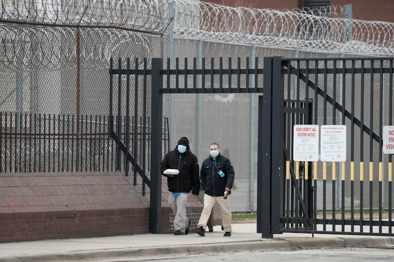 Two men with lanyard IDs and masks leaving a prison. The gate is surrounded by barbed wire.