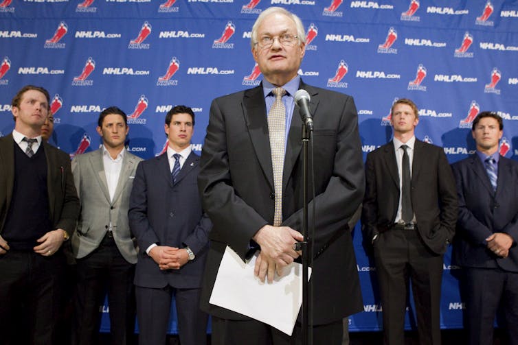 Man in a suit holding sheaf of papers stands in front of microphone. Five other men in suits stand in the background.