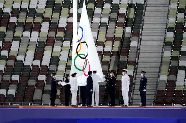 Masked officials stand on a stage as the Olympic flag is lowered, with empty seats in the background