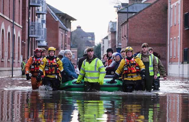 Rescue crews guide a large inflatable raft with several older residents seated inside through a flooded city street.