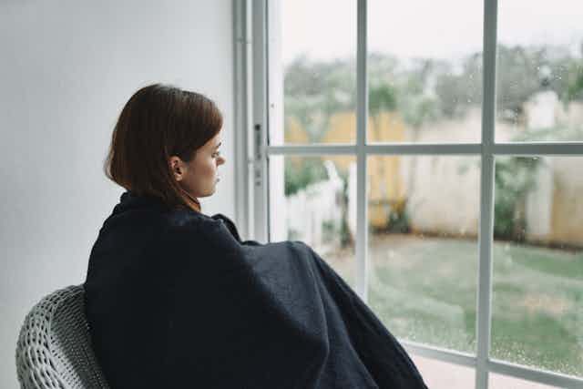 A young woman sits wrapped in a blanket looking out the window.