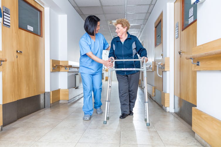 A nurse helps an older woman walk down a hospital hallway.