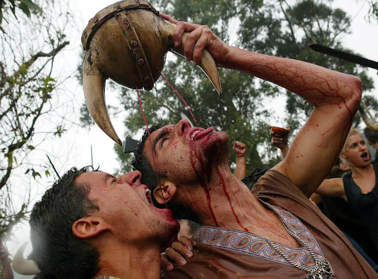 Two men drink wine from a Viking helmet.