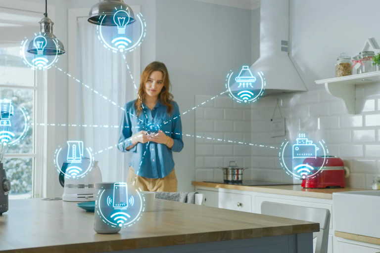 Une jeune femme debout dans une cuisine tient un téléphone intelligent relié à divers appareils électroniques.