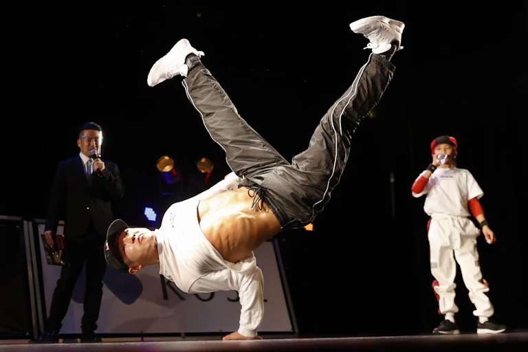 A man breakdances while a commentator and competitor look on.