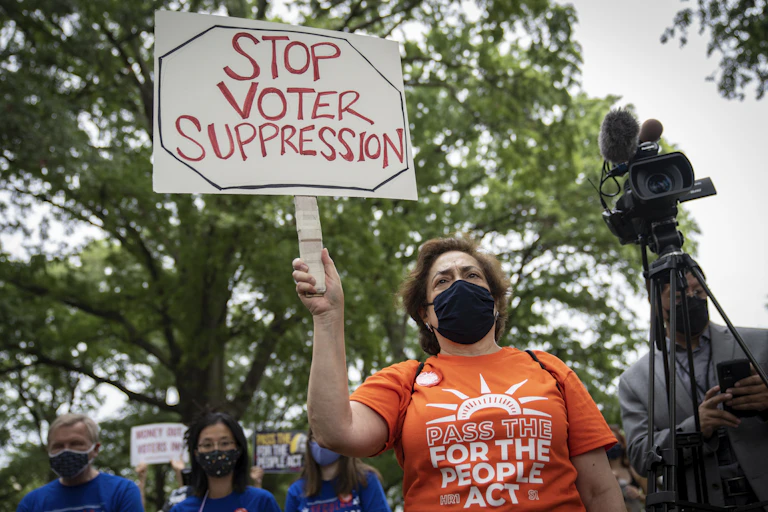 A woman in an orange shirt at a rally with a sign that says "Stop voter suppression"