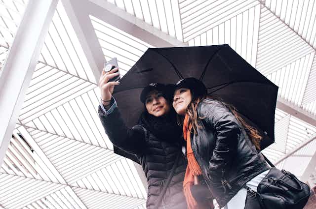 two women take a selfie photo with a smartphone while holding an umbrella and standing under a large roof-like structure with a distinct geometric pattern