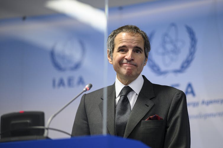 A man in a suit stands at a podium with the logo of the IAEA behind him.