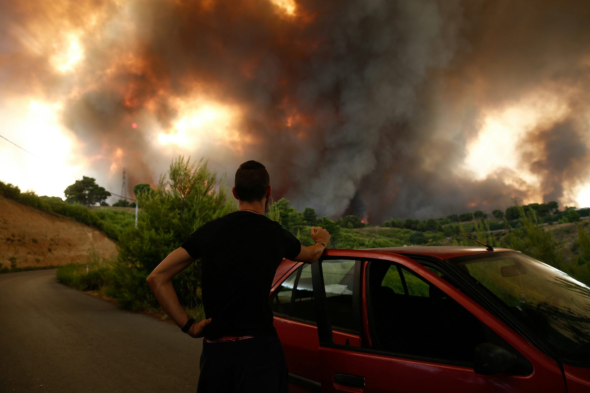 A man stands with his back to the camera, beside his car and watches smoke and fire billowing over the horizon in Greece.