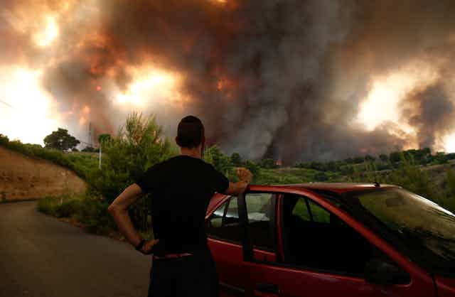 A man stands with his back to the camera, beside his car and watches smoke and fire billowing over the horizon in Greece.