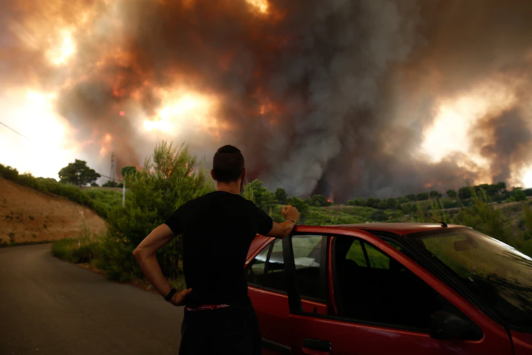 A man stands with his back to the camera, beside his car and watches smoke and fire billowing over the horizon in Greece.