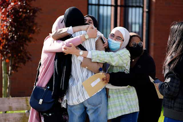 A group of students in headscarves and facemasks embrace with results sheets in hand