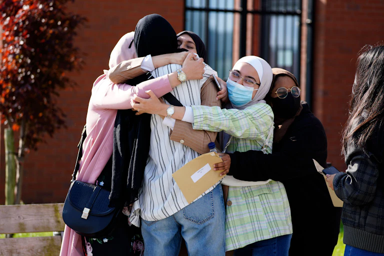 A group of students in headscarves and facemasks embrace with results sheets in hand