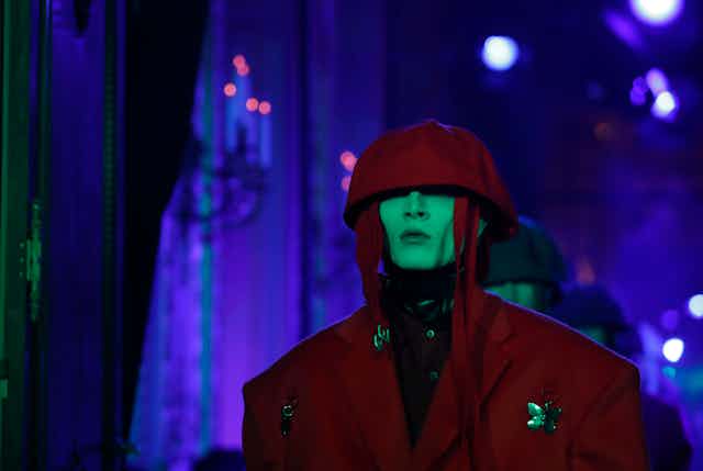 A male model with green makeup on his face wears a burgundy hooded jacket on a fashion runway.