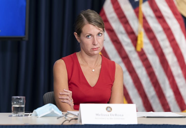DeRosa in a red dress sits with arms crossed and a stern, unhappy look on her face