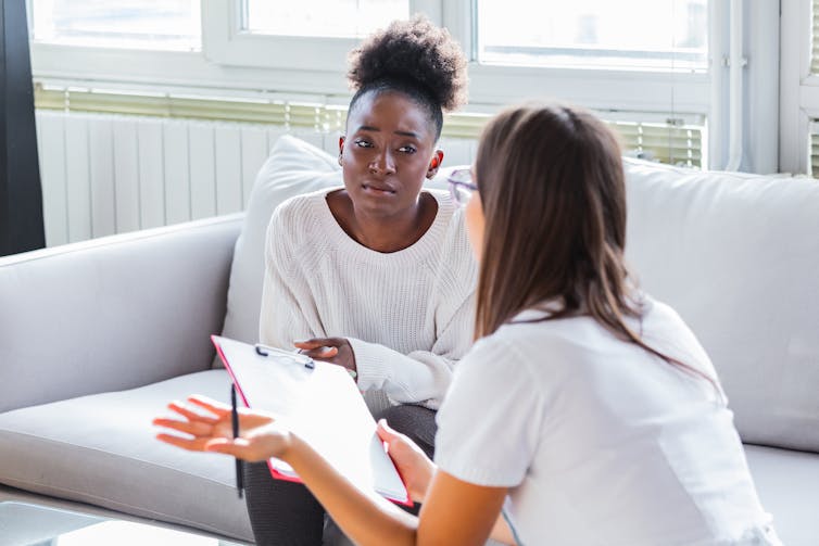 A woman sits on a couch as another woman speaks to her.