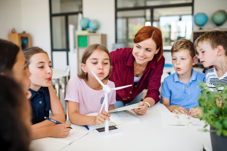 Female teacher and primary school students blowing a model wind turbine
