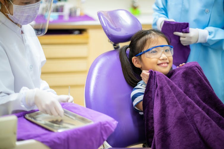 Child sits in a dentist's chair, holding a purple blanket to her chin.