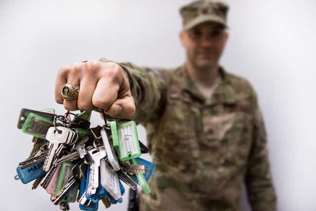 A man in a camouflage uniform extends his hand, holding many sets of keys