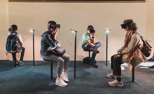Four people sit on stools in a museum room wearing virtual reality goggles