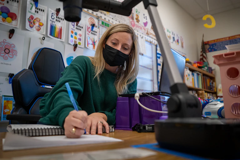 A teacher sits in a classroom in front of a laptop