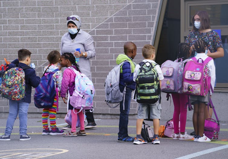 Teachers in face masks greet students in a schoolyard.