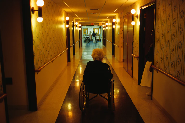 Older adult sitting in wheelchair going down the hallway of an assisted care facility.