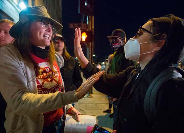 one unmasked woman argues with another who is wearing a mask, both holding up their hands