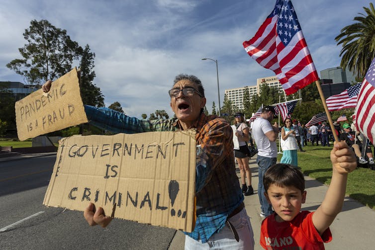 man with child holds homemade signs that read 'pandemic is a fraud' and 'government is criminal'