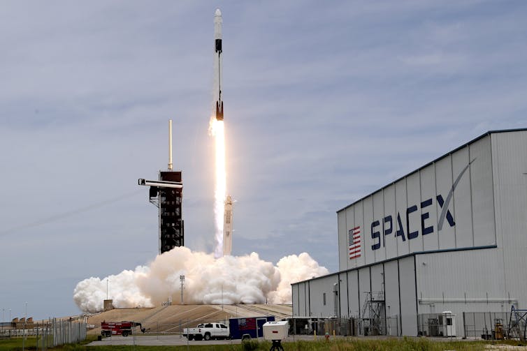 A rocket launches in the background, a SpaceX building in the foreground