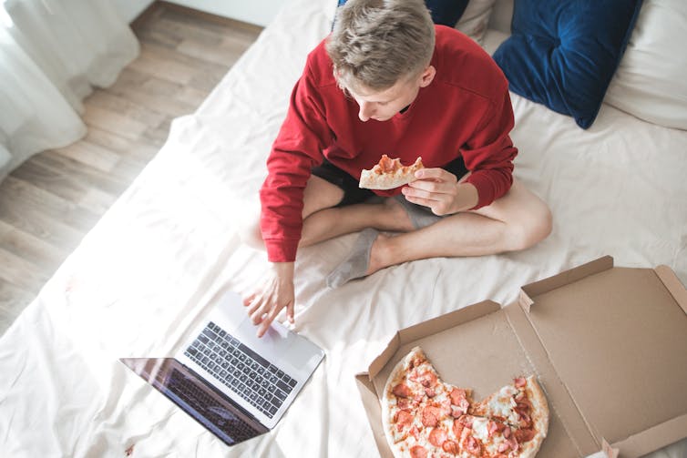 A young man sits on his bed eating pizza.