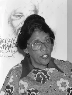 A woman in a floral jersey and glasses, hair in a bun, sits in front of a poster of Nelson Mandela.
