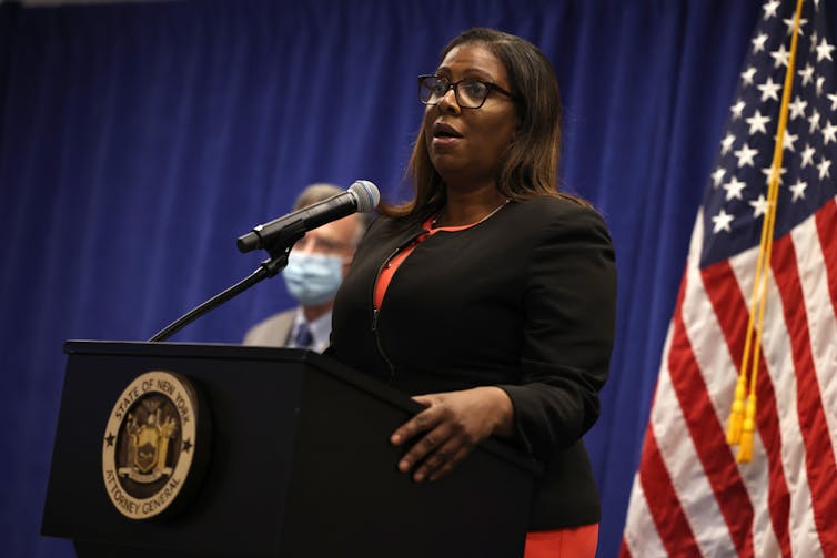 Letitia James, Attorney General of New York, standing in front of a lectern, speaking into a microphone, with an American flag behind her.