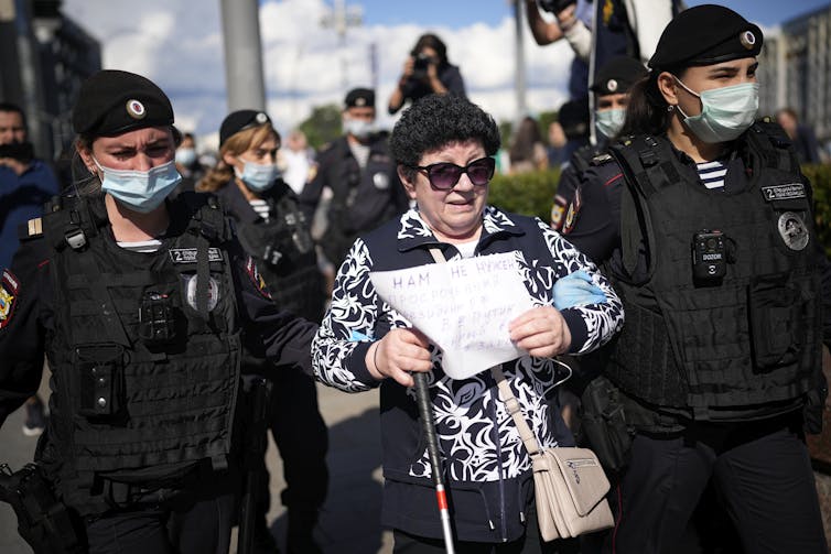 Woman is held on either side by police officers, she carries a sign at an anti-vaccination protest