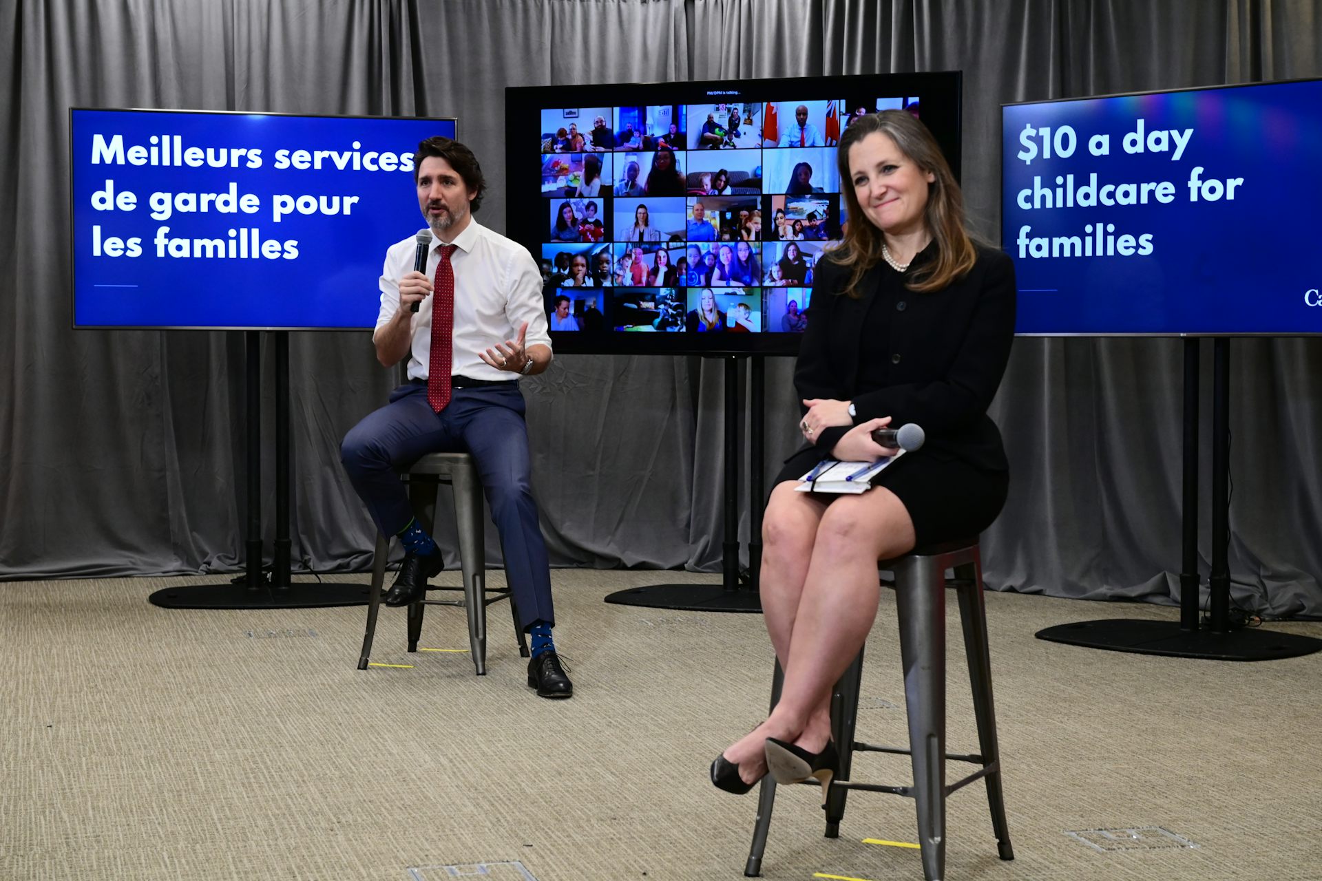Justin Trudeau and Chrystia Freeland sit in front of screens promoting the national child-care plan in both official languages.
