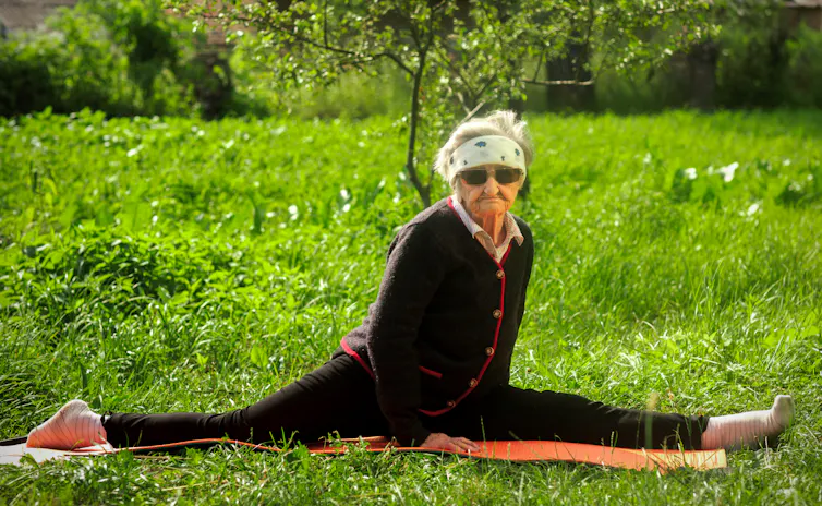 Photo d’une femme très âgée qui fait un grand écart dans son jardin, sur un tapis de sol.