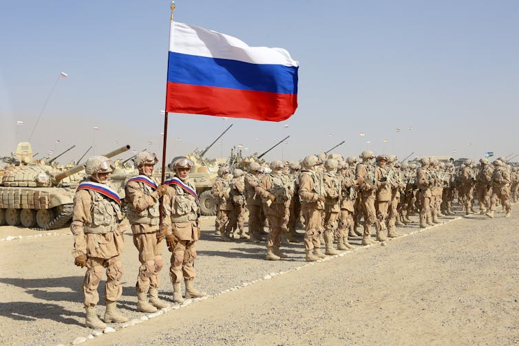 A group of soldiers stands alongside armored vehicles with a Russian flag flying overhead.