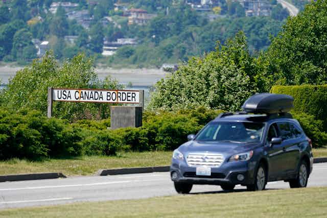 A subaru outback with roof racks, drives past a sign that reads 'USA CANADA BORDER'