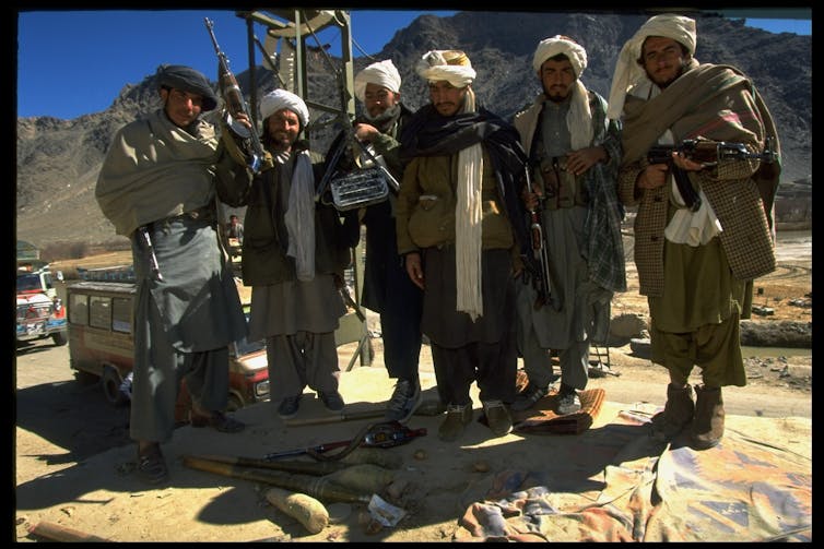 Six armed men in head coverings with beards stand by a road with mountains in background.