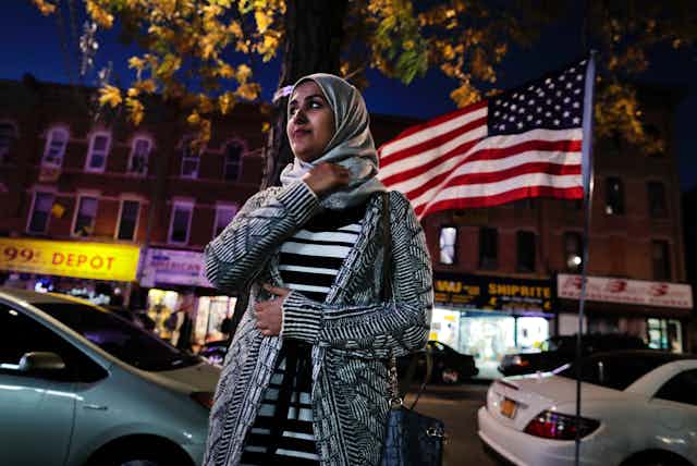 A young woman wearing a headscarf stands on a street in front of an American flag