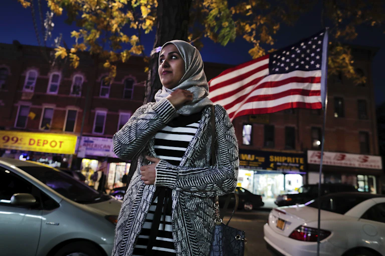 A young woman wearing a headscarf stands on a street in front of an American flag